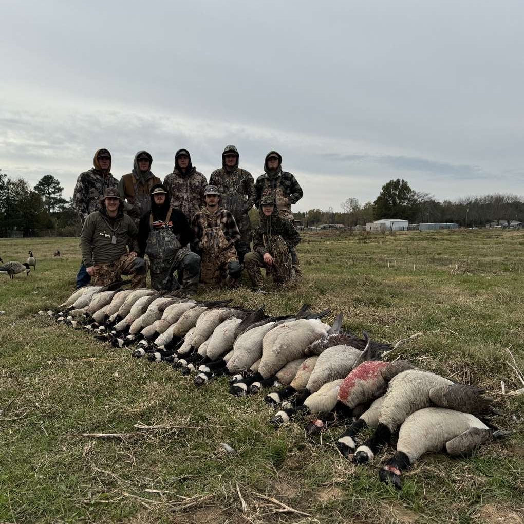 Group of people in camouflage posing with a row of hunted animals on a grassy field.